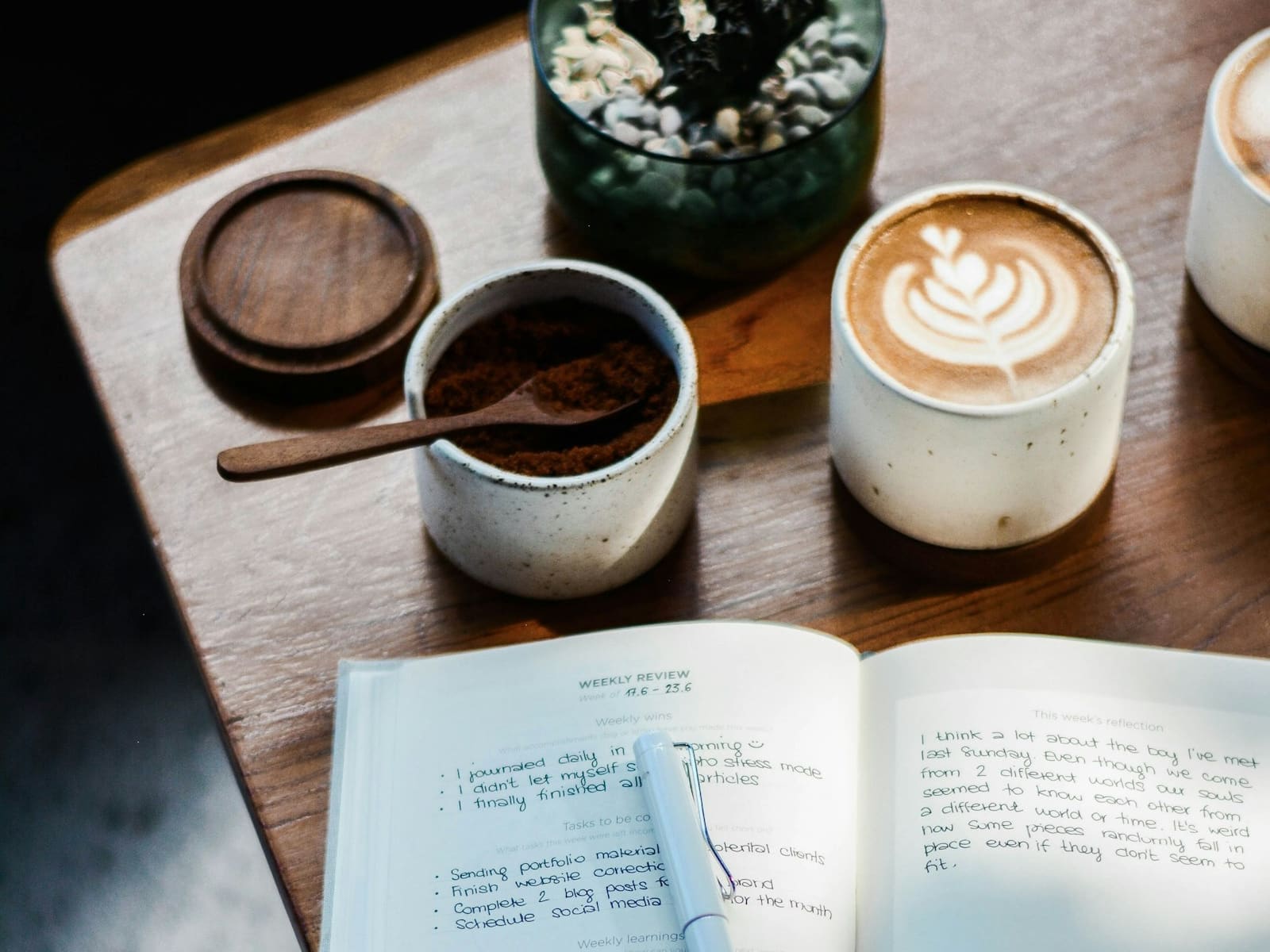 Offenes handgeschriebenes Notizbuch neben Kaffee und Pflanzen auf einem warmen Holztisch